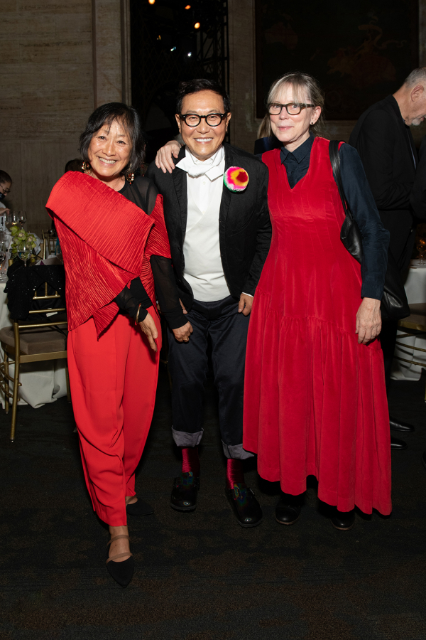 Color photograph of three people at a fundraising gala smiling at the camera: an Asian woman and a light skinned woman, both wearing fancy and stylish black and red dresses flank an Asian man wearing formal wear but with his pants cuffed rolled up