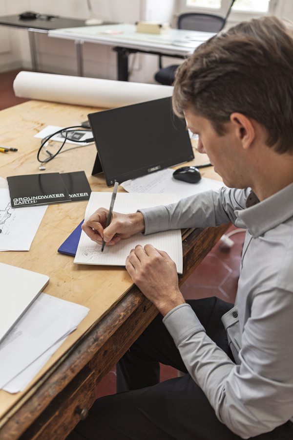 Color photo of a light skinned man seated in a chair at a desk, writing or drawing in a notebook