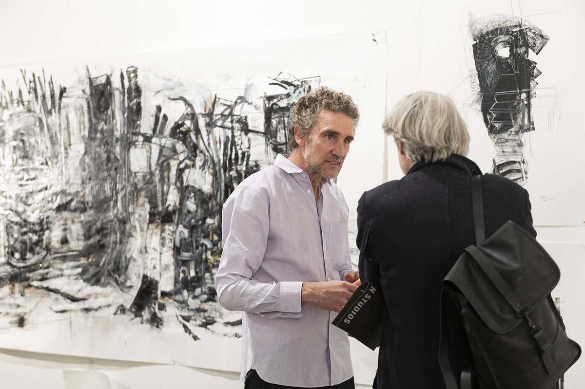 Color photo of a man talking to another man in an artist's studio with drawings on the wall behind them