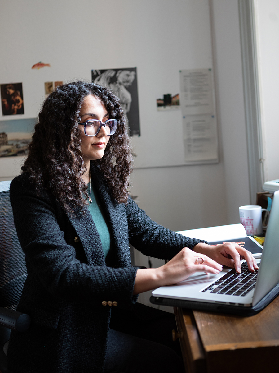 color photo of a young woman sitting at a desk in a white-walled study, typing on her open laptop