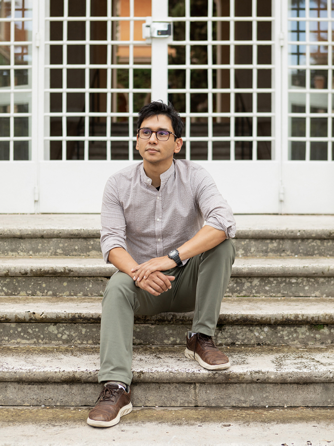 Color photo of an olive skinned man sitting on stone steps in front of a gridded white gate