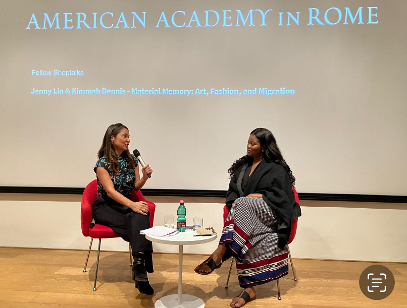 Color photo of two women in chairs at the front of a lecture room, talking to each other