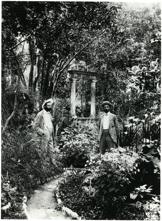 black and white photo of two men standing in a lush garden; the photo was taken over 100 years ago