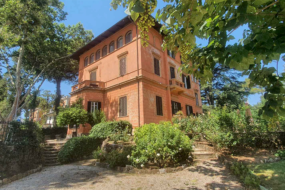 Color photo of a red-walled two-story villa in Rome, surrounded by trees and bushes