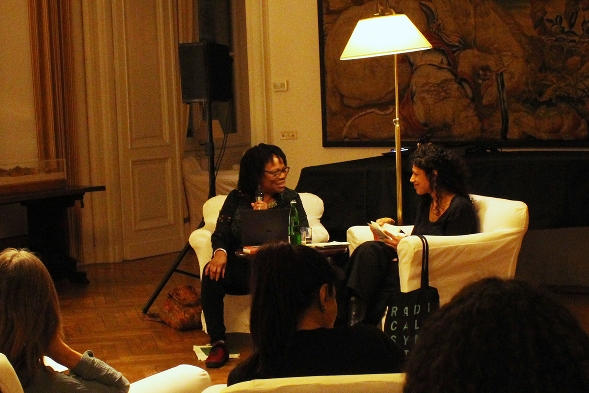Color photo of two Black women sitting in chairs in a living room setting and talking to each other with microphones
