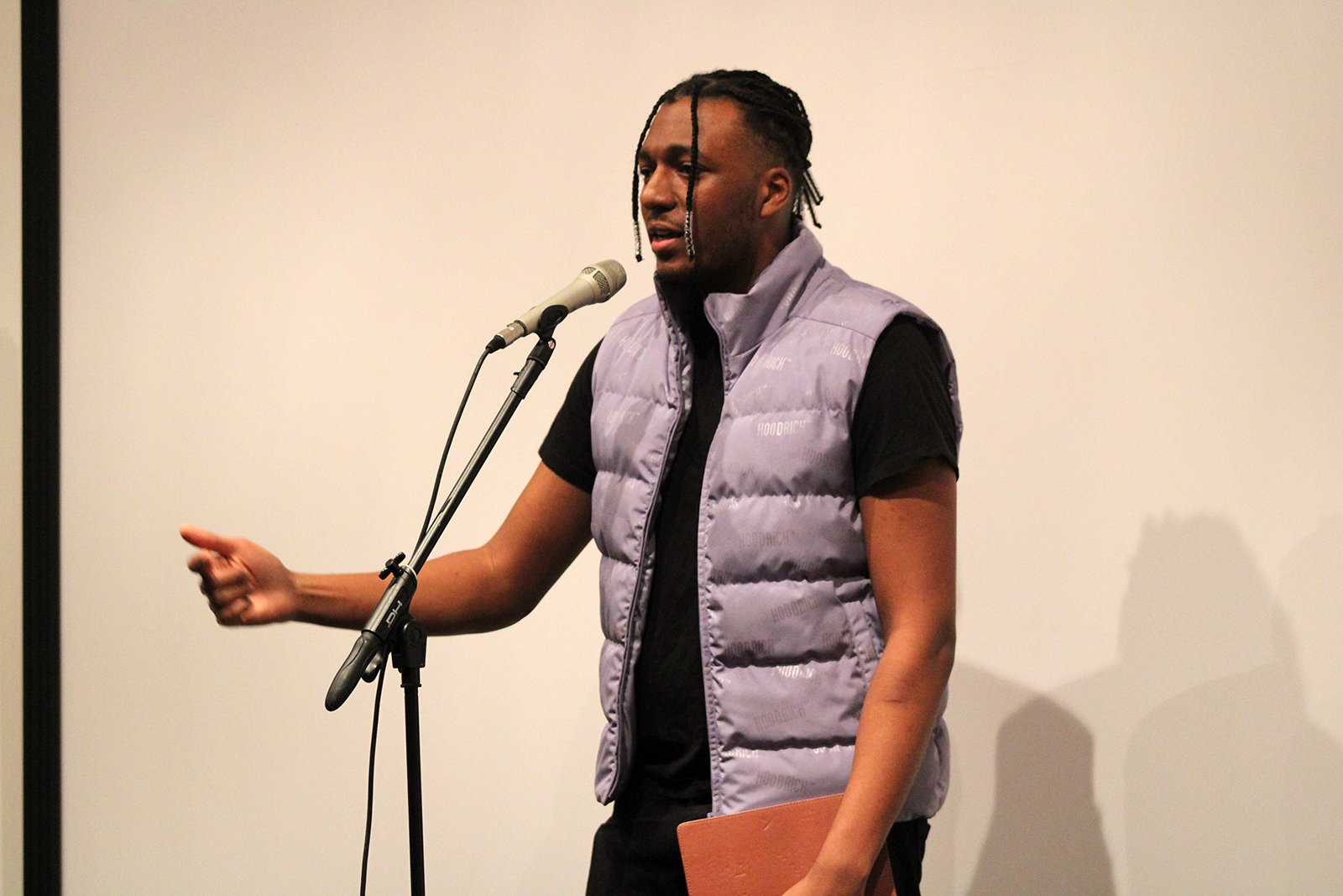 Color photo of a Black man wearing a lavender puffer vest and standing in front of a microphone and a white wall