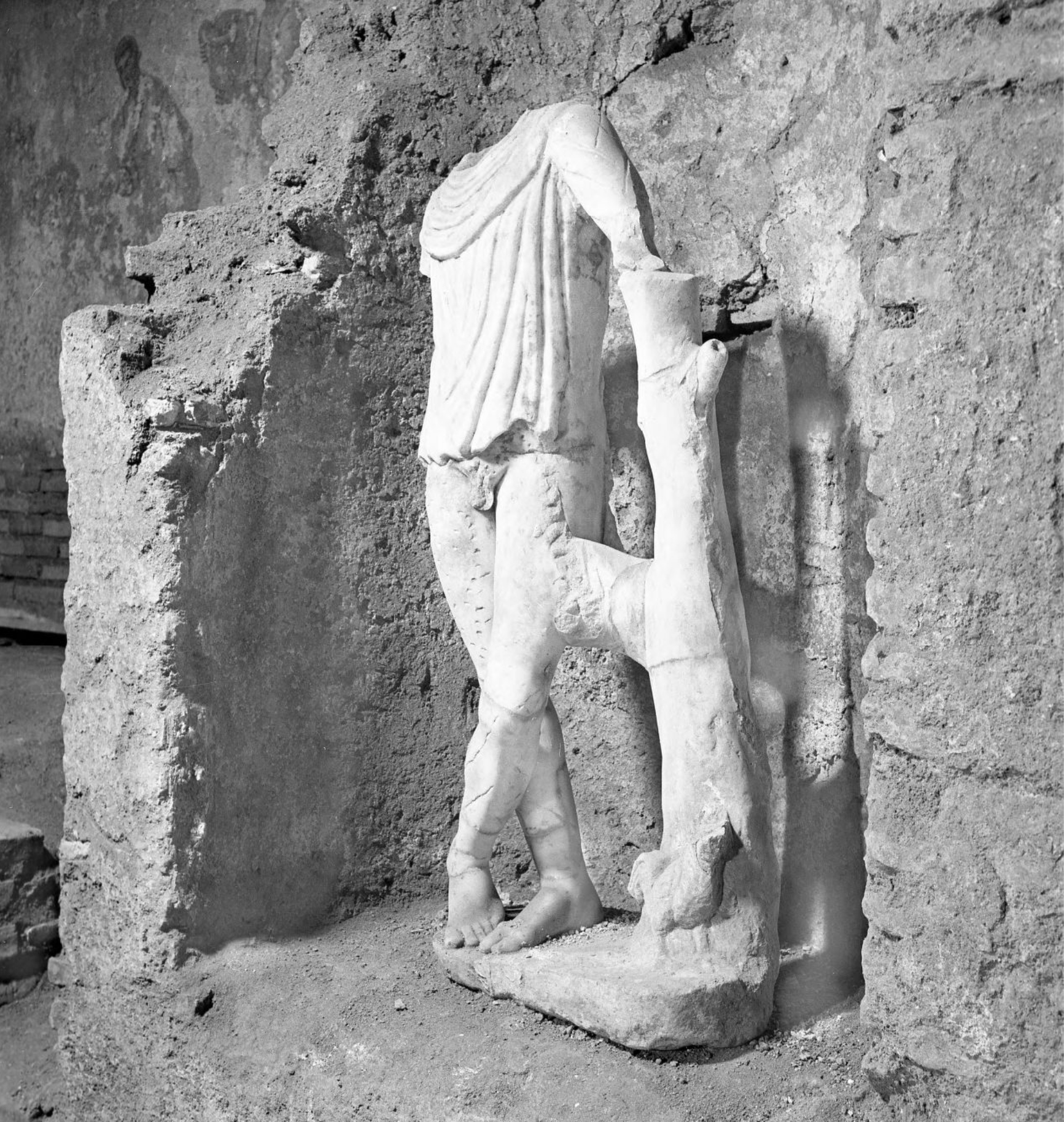 Black and white photograph of an ancient Roman statue with a missing head arms; the work is positioned in a niche in a stone wall
