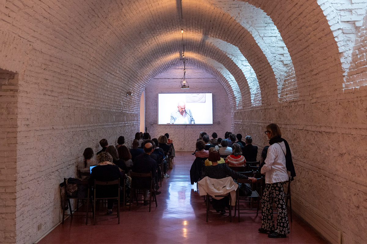 color photo of people seated in chairs in front of a video screen projecting a lecture on the wall of a cryptoporticus