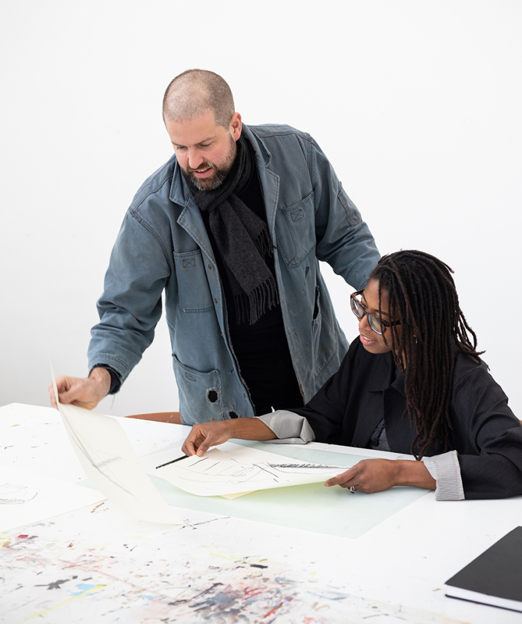 Color photo of a white man and a black woman at a work table looking at drawings in front of them