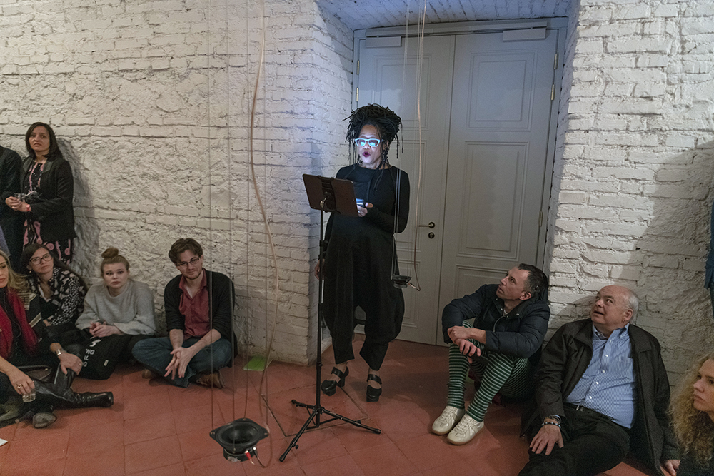 Color photograph of Pamela Z performing a sound art installation in the Cryptoporticus of the American Academy in Rome; she is standing in front of a music stand, and audience members are sitting on the ground to her left and right