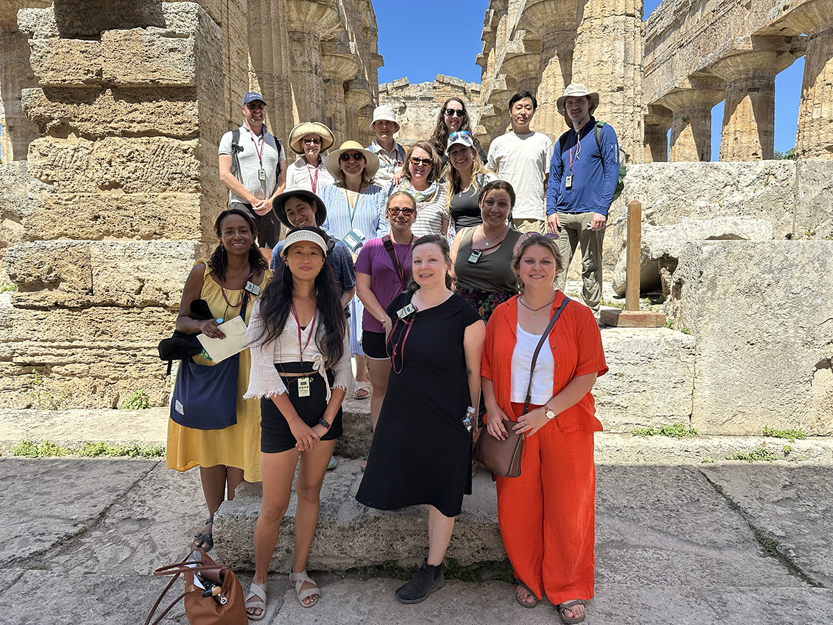 color photo of 16 people posing for a group portrait while standing in front of Romann ruins on a sunny day
