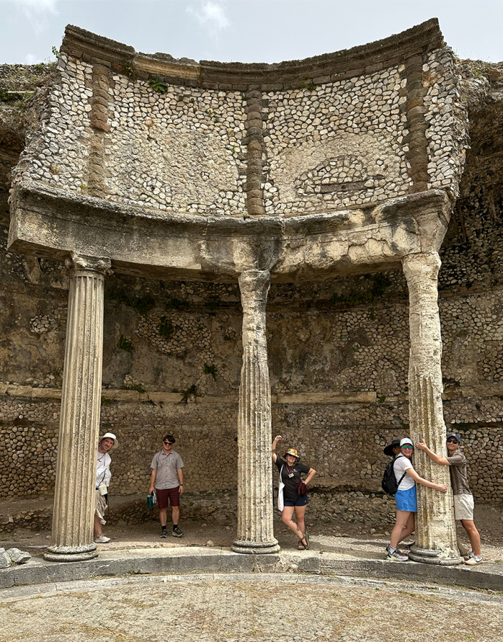 Color photo of 5 people standing in front of the ruins of an ancient roman building; two people are hugging a column