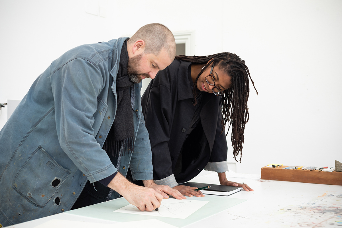 Color photograph of a light skinned man and dark skinned woman, looking at papers on a worktable in front of them
