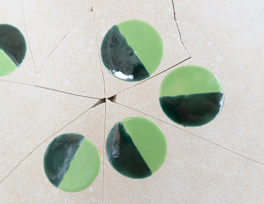 Color photograph with a close-up view of a wall-mounted artwork consisting of glazed and cracked off-white-colored ceramic tiles with painted green circles
