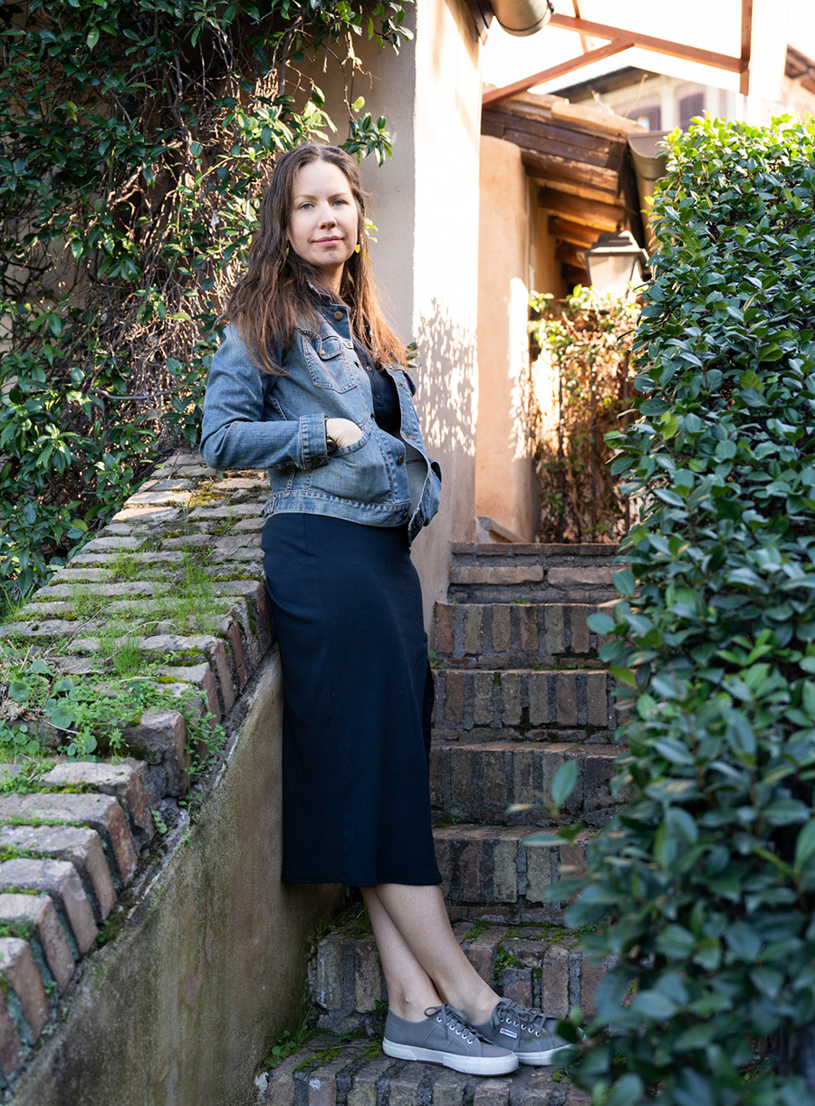Color photo of a light skinned woman wearing blue jacket skirt and shoes standing on brick staircase to a building in a garden