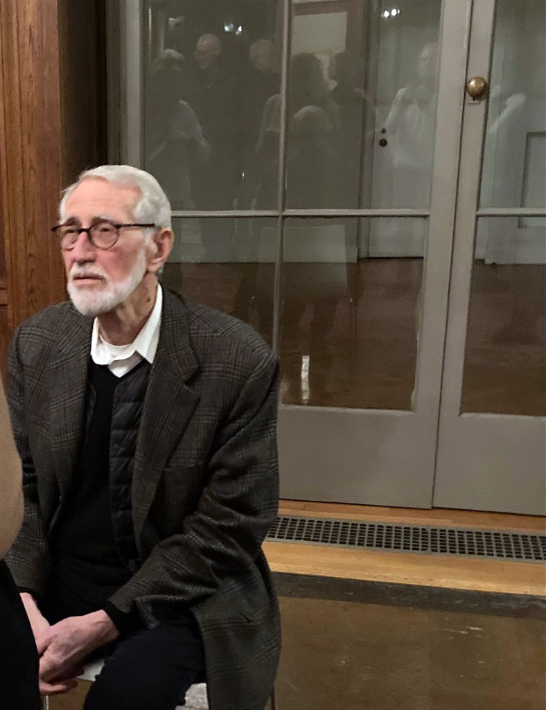 Color photograph of a light skinned man wearing glasses and a white beard sitting in a chair