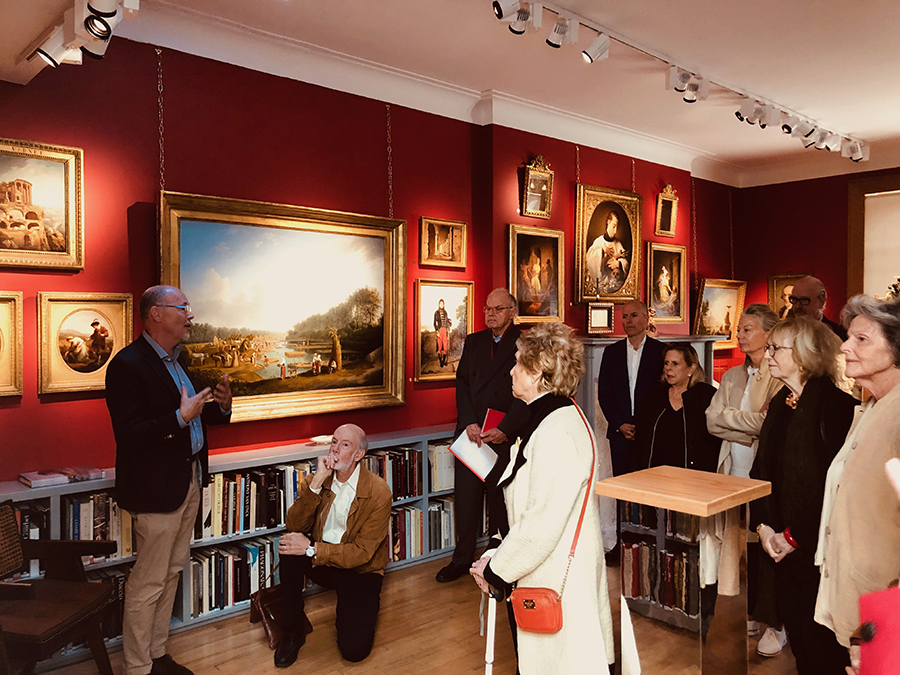 Color photograph of a man leading a tour of about ten people in a gallery with various paintings hanging from red walls