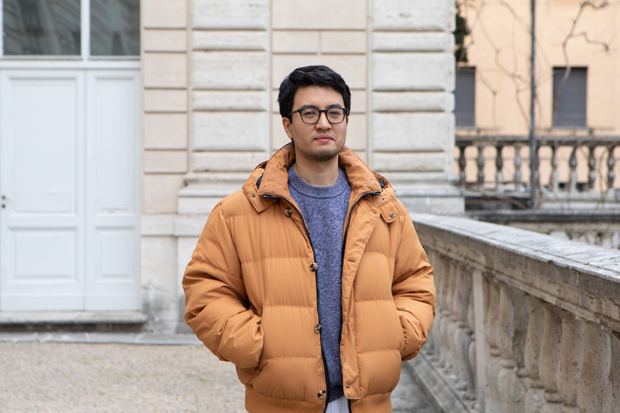 Color photograph of an Asian man wearing glasses and an earthy orange colored puffer coat; he stands outside the main entry doors of the American Academy in Rome with his hands in his jacket pockets