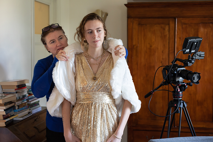 Color photograph of an older light skinned woman placing a fur coat over a younger light skinned woman; to their left is a movie camera on a tripod