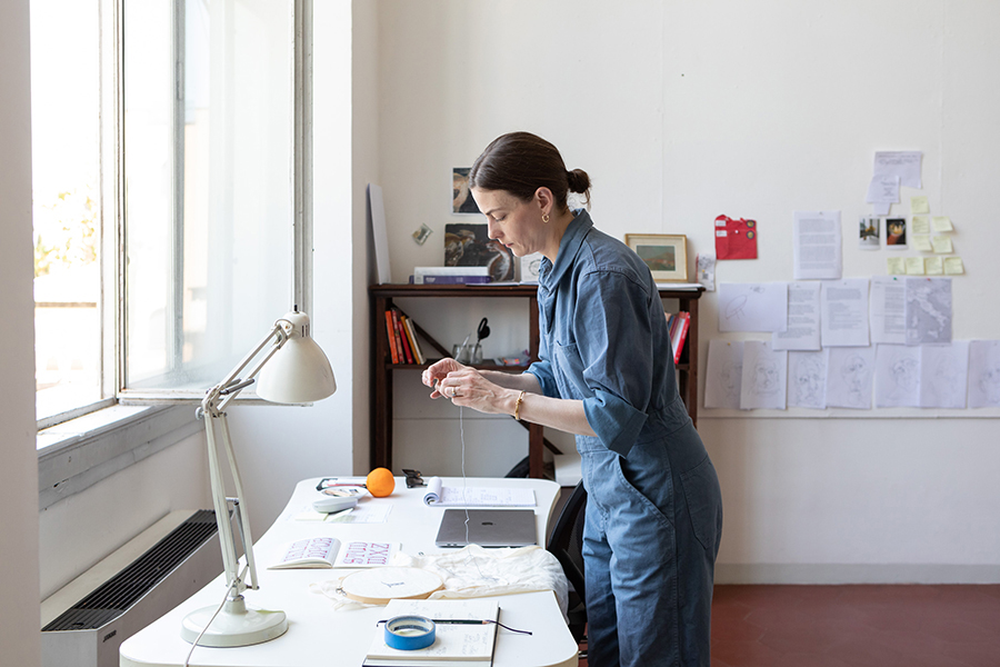 Color photograph of a light skinned woman wearing a blue jumpsuit in profile; she is standing in front of a desk in her studio and sewing with thread