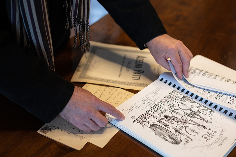 Color photograph of the hands of a light skinned man holding a printed musical score; on the table in front of him are other historical printed documents on paper