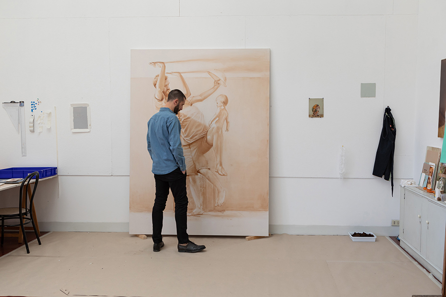 Color photograph of the artist Manuele Cerutti in his studio, standing in front of and looking at his large painting