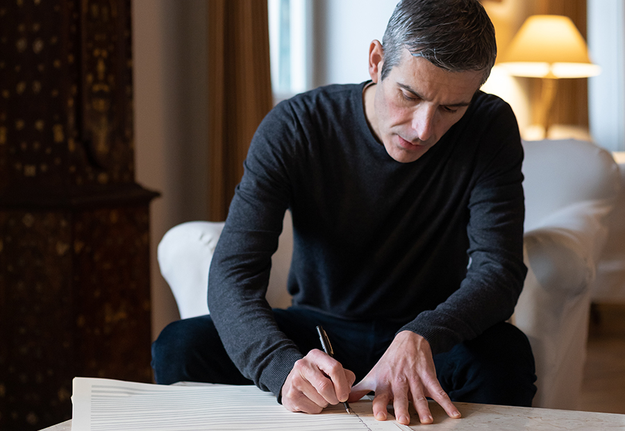 Color photograph of a light skinned man sitting in a room; he is writing on a large musical score on a low table in front of him