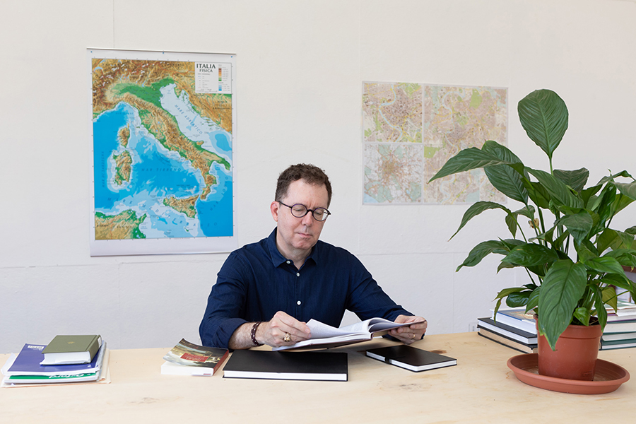 A light skinned man wearing glasses sits at a table reading a book. Also on the table are more books and a medium sized potted plant. Behind the man (Michael Lee) are maps of Italy and Italian cities