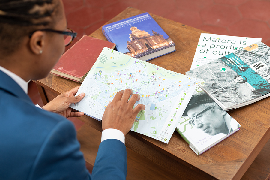 Color photograph of the back of the head and shoulder of a dark skinned woman with dreadlocks; she holds a map of Rome in her hands and books are strewn about the table in front of her
