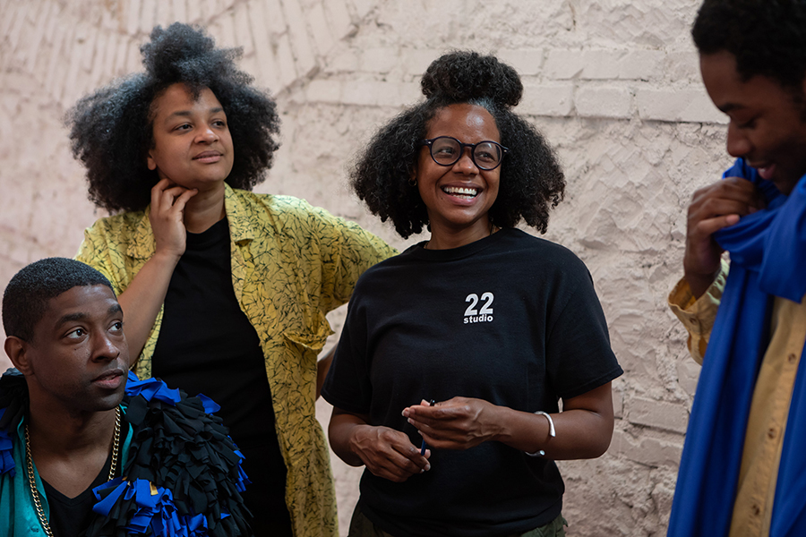 Color photograph of two dark skinned women (the artists known as Las Nietas de Nonó) smiling and looking to their left at a dark skinned man; a fourth black man sits to the artist's right