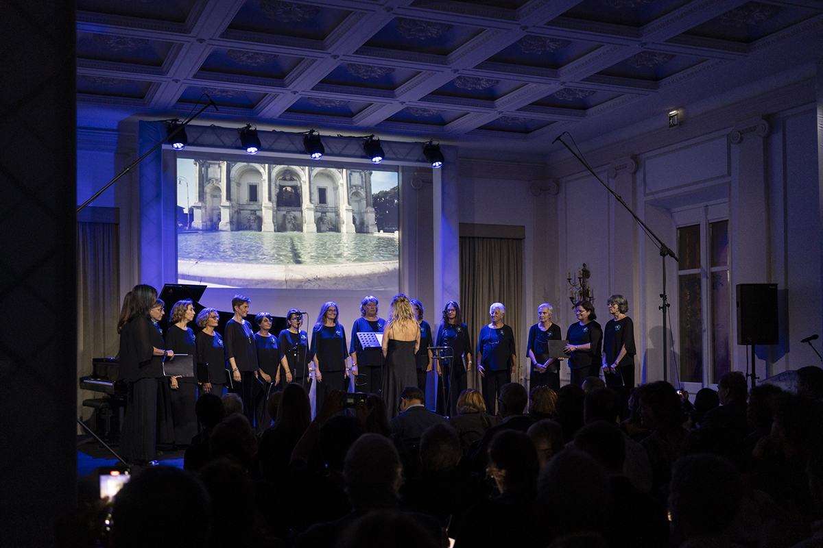 Color photograph of an all-female choir standing onstage with a photograph of a large Roman fountain projected on the wall behind them