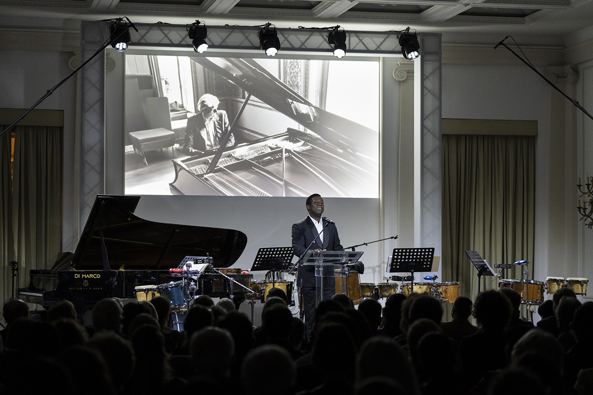 A dark skinned man stands at a podium overlooking an audience with a photograph of a pianist projected on the wall; behind him is a piano, several sheet music stands, and various percussion instruments