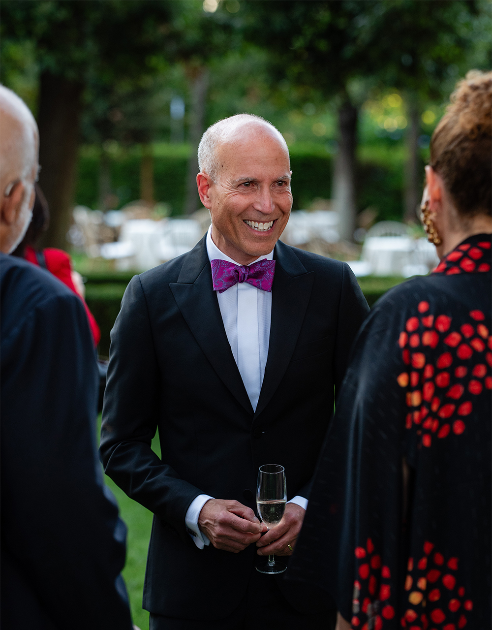 Color photo of a light skinned man wearing a dinner jacket and pink purple bowtie talks to an unseen woman in a garden location