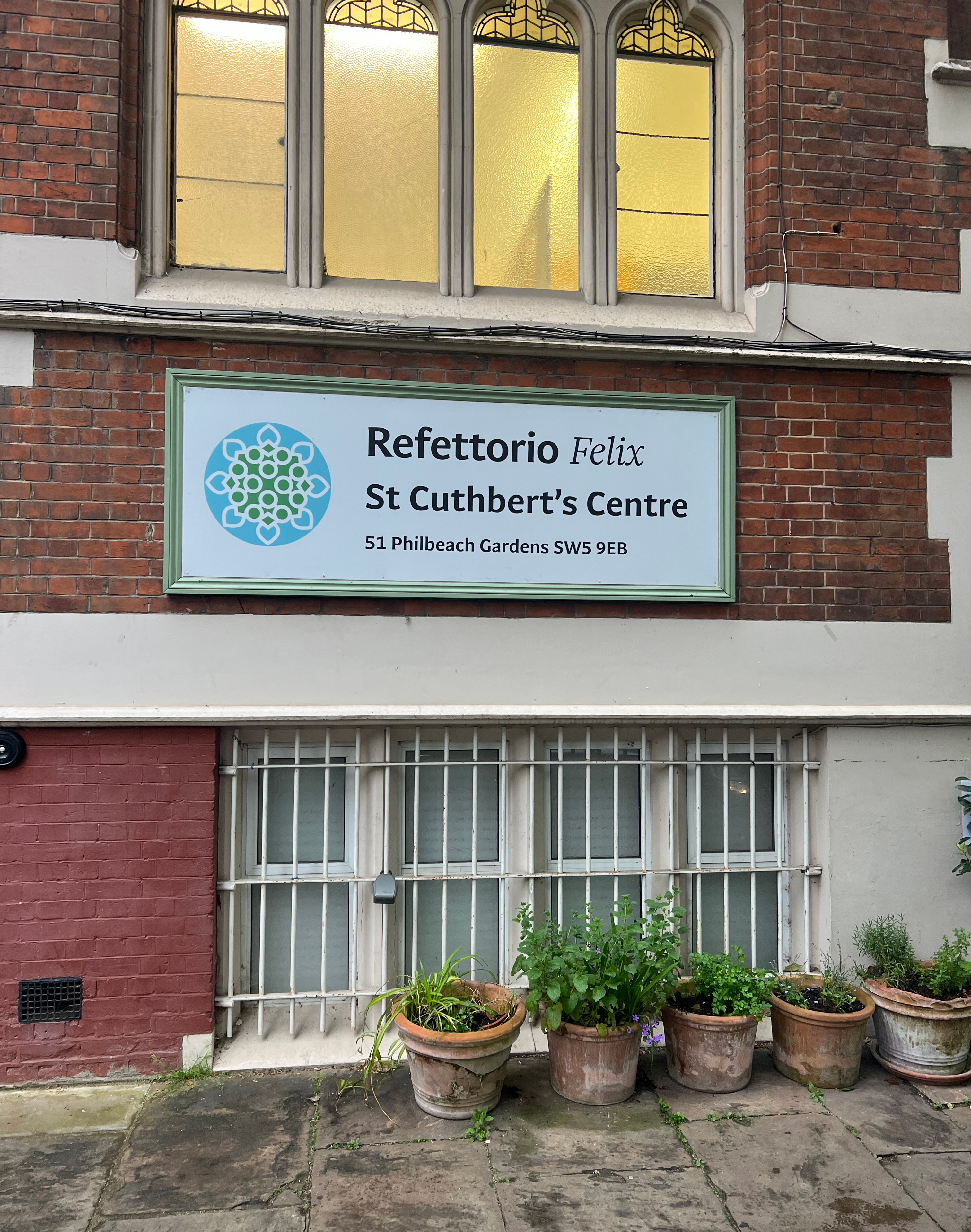 Color photo of an exterior wall and window of a brick building in London, with five potted plants on the sidewalk