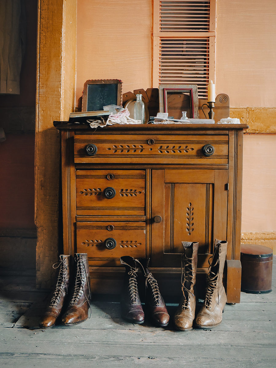 Color photograph of a historical museum exhibit with three pairs of nineteenth century lace-up boots on the floor in front of a small dresser or cabinet