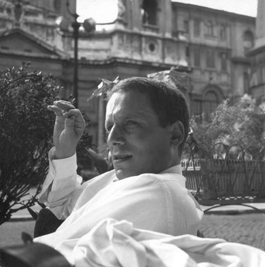 Black and white photograph of the head, shoulders, and right hand of a light skinned man sitting at a cafe table in the Piazza Navona, with baroque buildings behind him