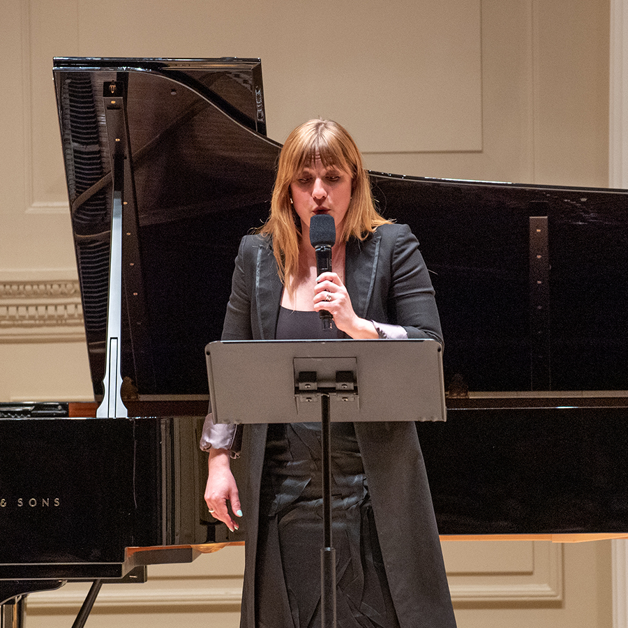 Color photo of a light skinned woman singing onstage holding a microphone, with a grand piano behind her