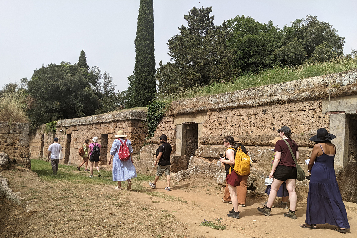 Color photo of a line of people walking outdoors on a dirt path alongside an ancient Roman wall