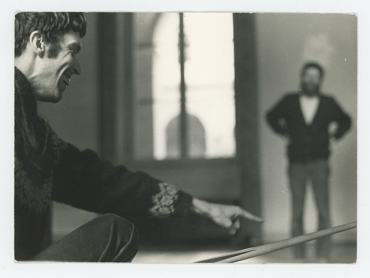 Black and white photo of a light skinned man kneeling and smiling; away from him and out of focus is a standing light skinned man