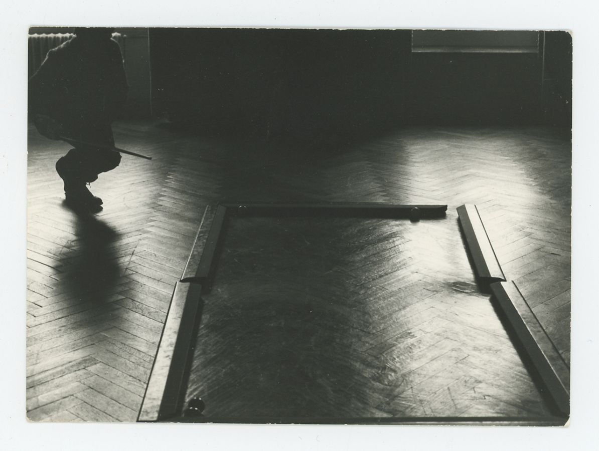 Black and white photo of a darkened room with wood slat floors; a person with a pool cue approaches a billiards "table" on the floor without a tabletop or legs