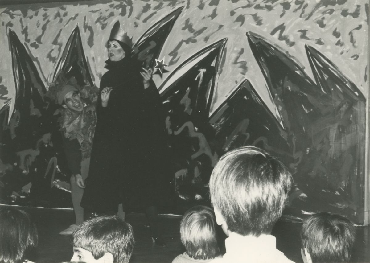 Black and white photo of two performers (man and woman) in costume standing in front of an abstract painted backdrop