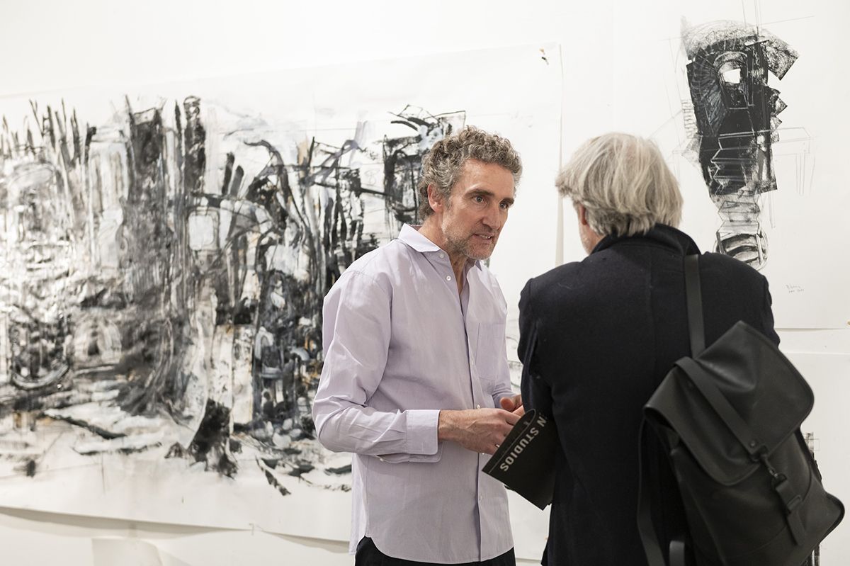 Color photo of a man talking to another man in an artist's studio with drawings on the wall behind them