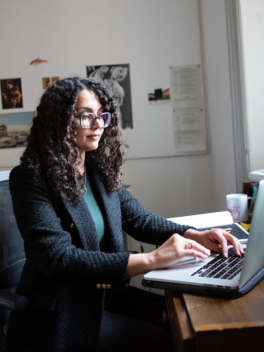 color photo of a young woman sitting at a desk in a white-walled study, typing on her open laptop