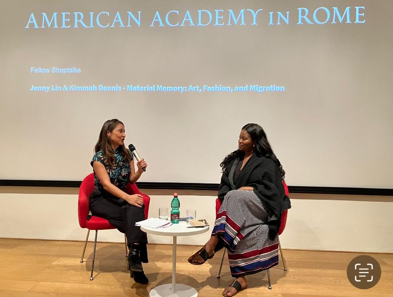 Color photo of two women in chairs at the front of a lecture room, talking to each other