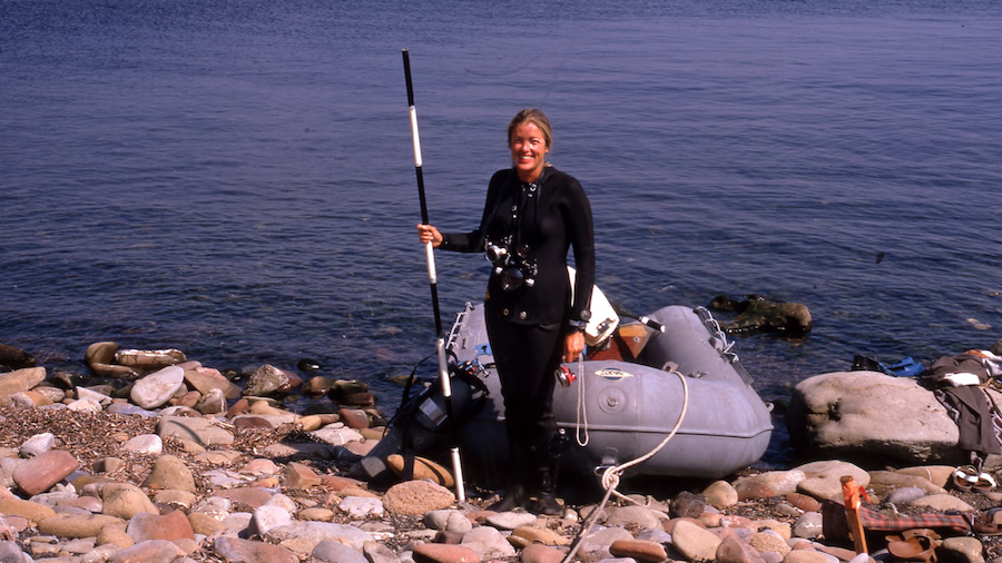 A woman holding a pole next to a dinghy, the sea behind