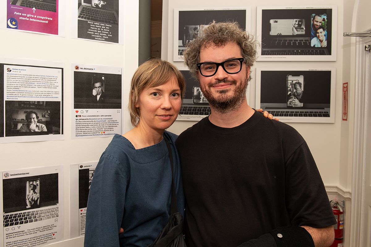 Color photo of a light skinned man and woman smiling at the camera standing in front of computer printouts in an art installation