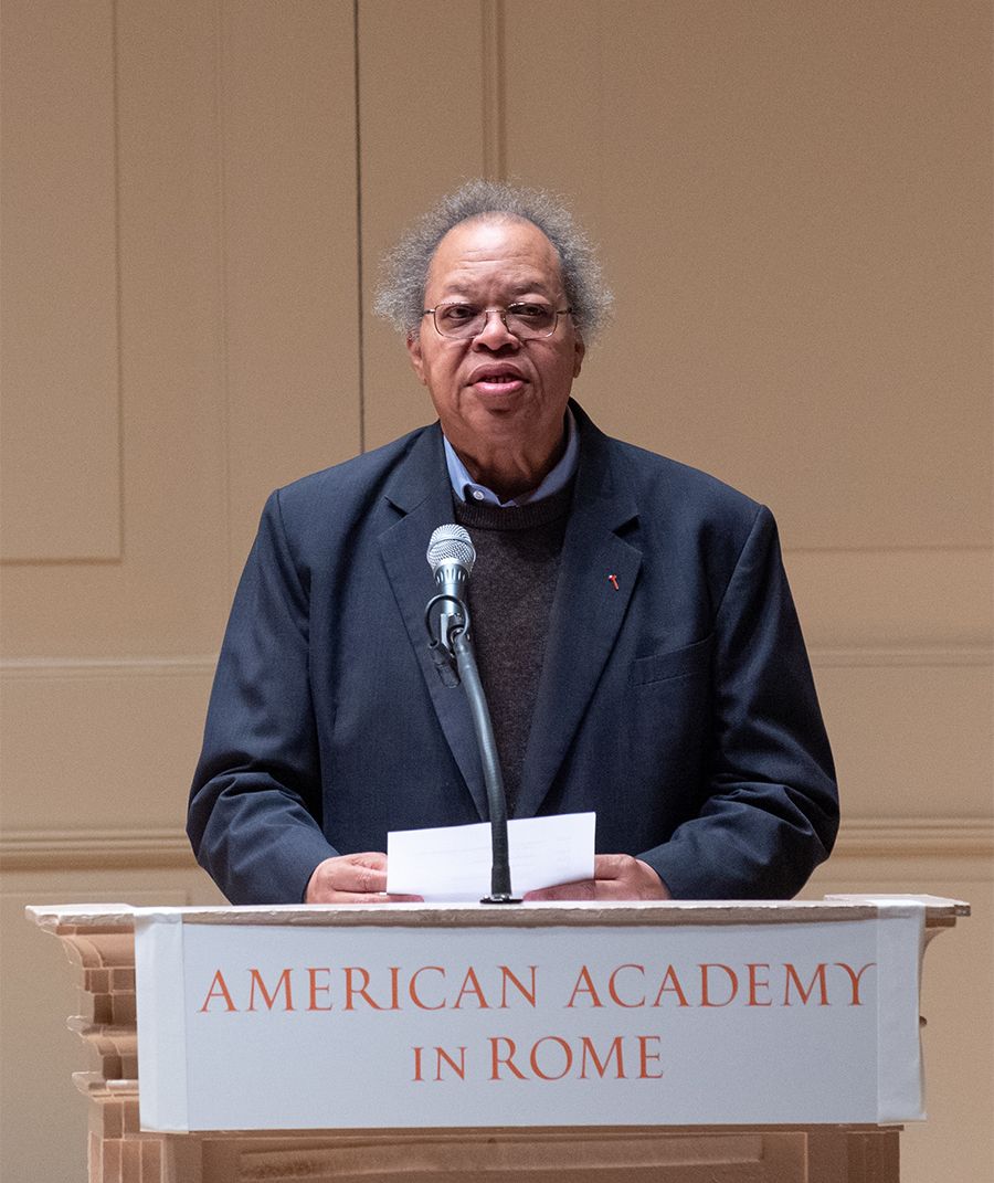 Color photo of a darker skinned man speaking at a podium in a concert hall