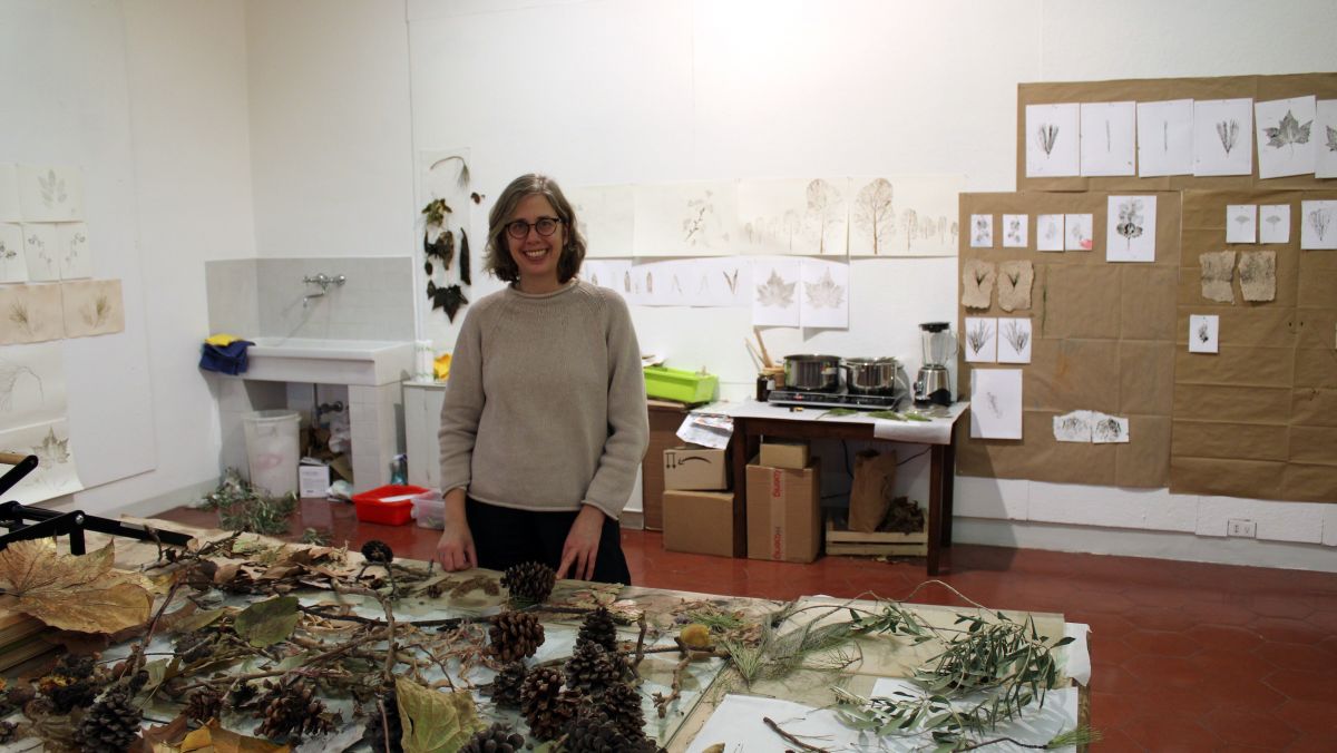 color photo of a light skinned woman in an artist's studio with botanical art on the wall behind her