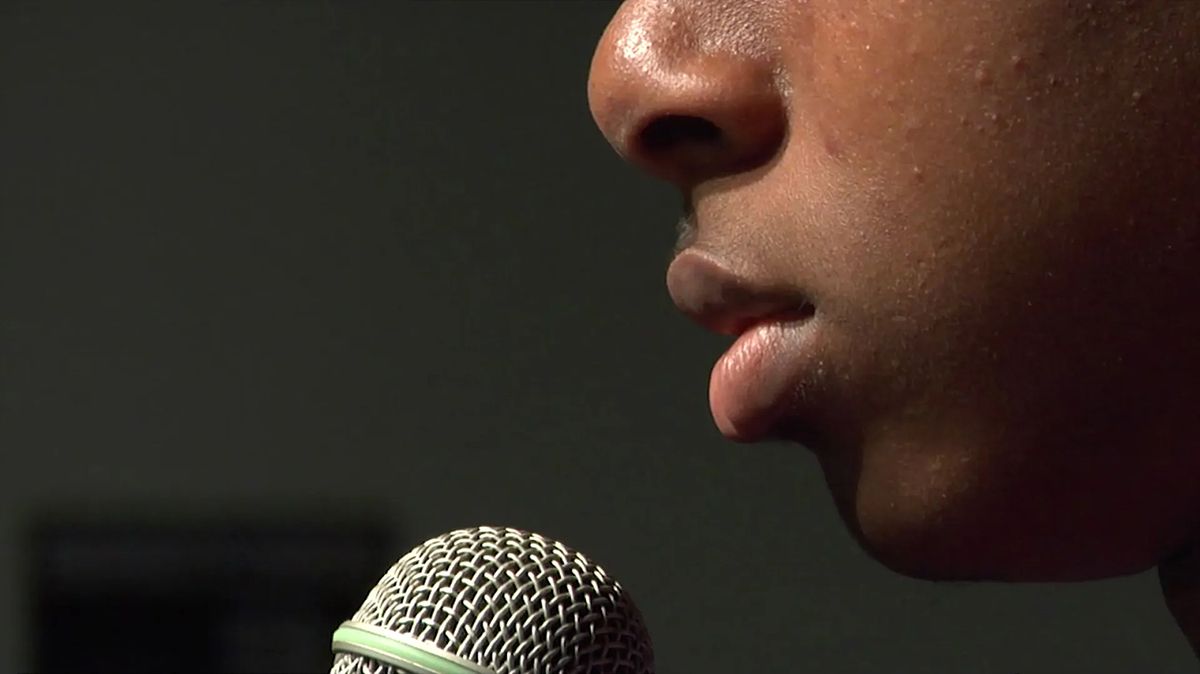 Color still from a video showing a close up of a dark skinned man's face in front of a microphone