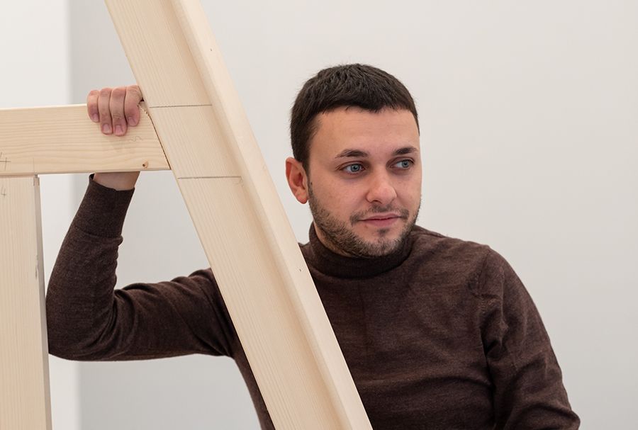 Color photograph of a light skinned man wearing a brown turtleneck looking off camera; he stands next to a wooden scaffold